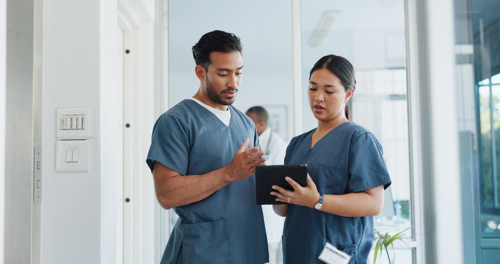 Two clinicians in scrubs talking over a tablet in a hospital corridor