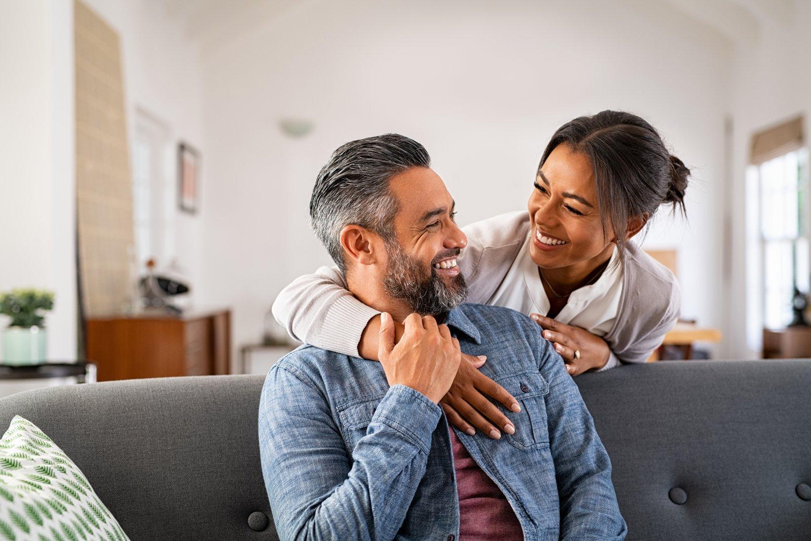 A smiling couple at home, one hugging the other from behind