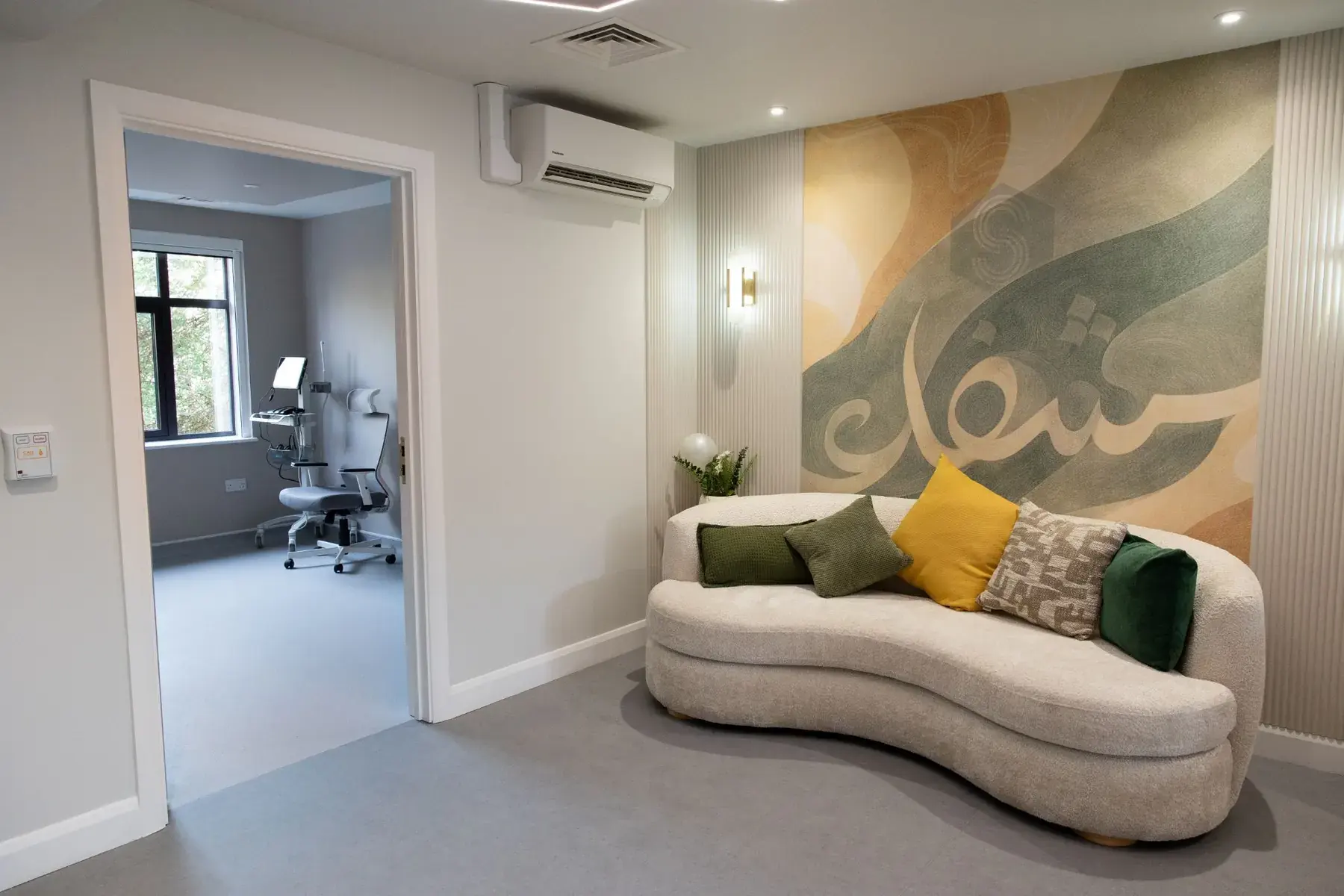 A curved sofa with green and yellow cushions beneath a calligraphy mural, beside an open door into a consulting room